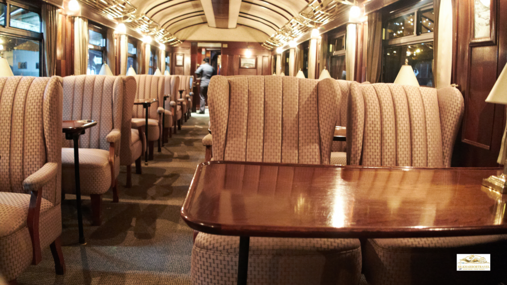 Wood-stained luxury train interior with polished tables, upholstered seats, and warm ambient lighting.