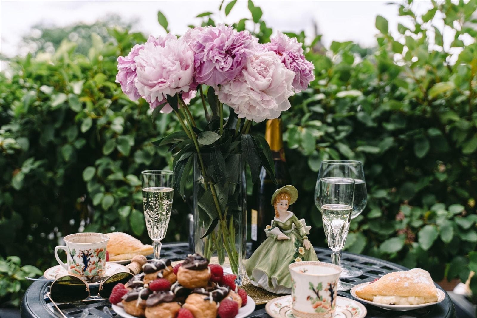 An outdoor table laid with china teacups, champagne flutes, pastries and peonies
