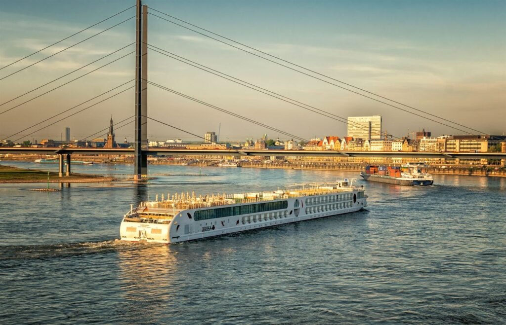 A long, low river cruise vessel moving along a river past a European city bridge and waterfront